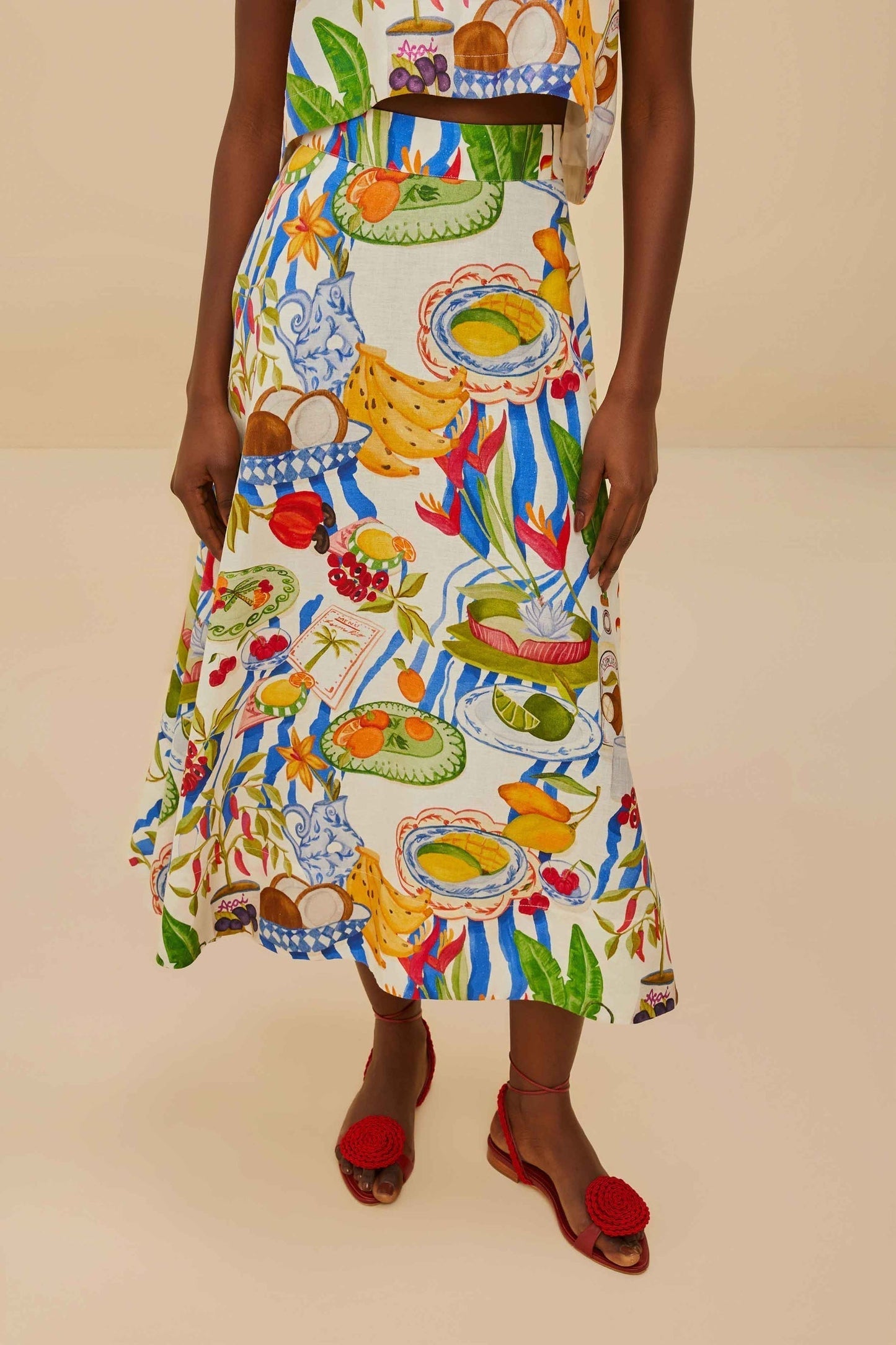 Colorful - Woman wearing a vibrant two-piece co-ord set featuring a sleeveless crop top and high-waisted A-line midi skirt, adorned with a colourful tropical fruit and plate print.