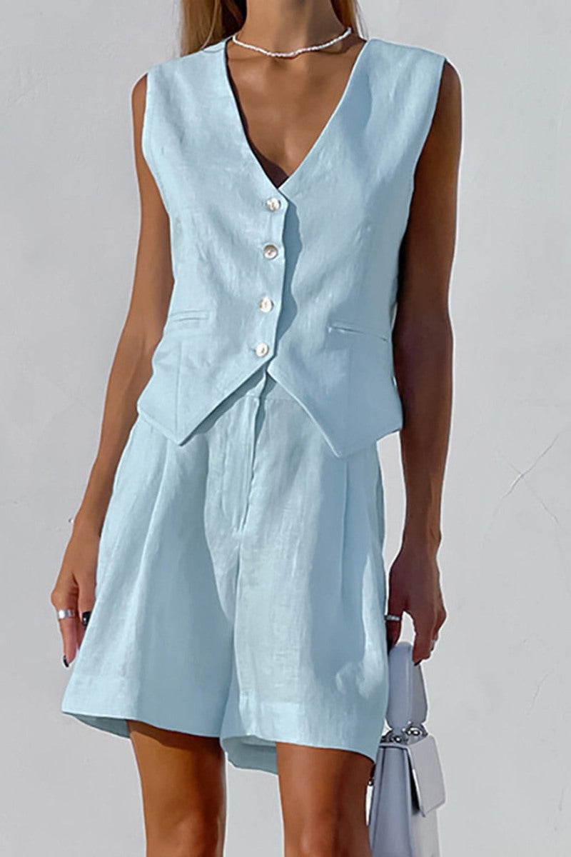 Blue - A woman wearing a white, sleeveless, button-front tailored waistcoat top paired with matching high-waisted pleated shorts, standing against a textured white wall.