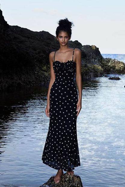 Black - A woman standing by the water wearing a black maxi dress with white polka dots, featuring spaghetti straps and a ruched bodice.