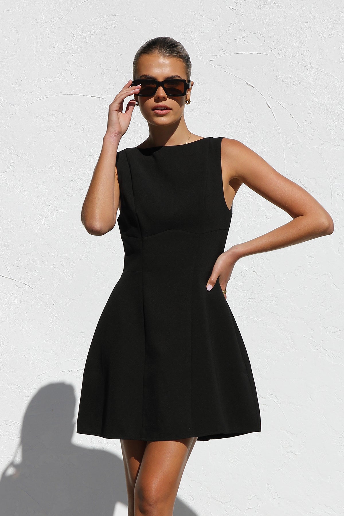 Black - A woman with blonde hair in a bun, wearing black rectangular sunglasses and a sleeveless, black, A-line mini dress with bodice seams, stands against a white textured wall in bright sunlight.