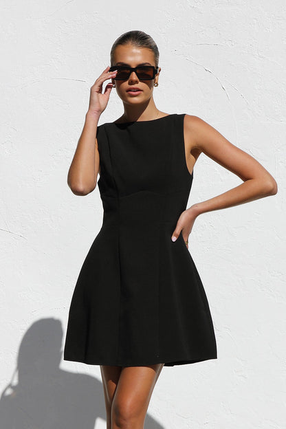 Black - A woman with blonde hair in a bun, wearing black rectangular sunglasses and a sleeveless, black, A-line mini dress with bodice seams, stands against a white textured wall in bright sunlight.