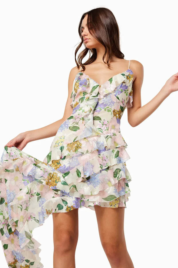 Green - A woman wearing a floor-length, ruffled maxi dress with a pastel floral print on a sheer cream background, featuring spaghetti straps and an asymmetric hem.