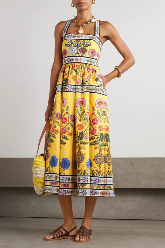 Woman wearing a bright yellow halter midi dress featuring a vibrant botanical floral print and contrasting geometric borders, styled with a woven tote bag and brown sandals.