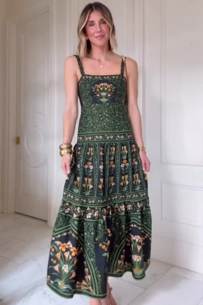 Woman wearing a dark green and black maxi dress featuring a square neckline, wide straps, a tiered skirt, and an intricate botanical floral border print.