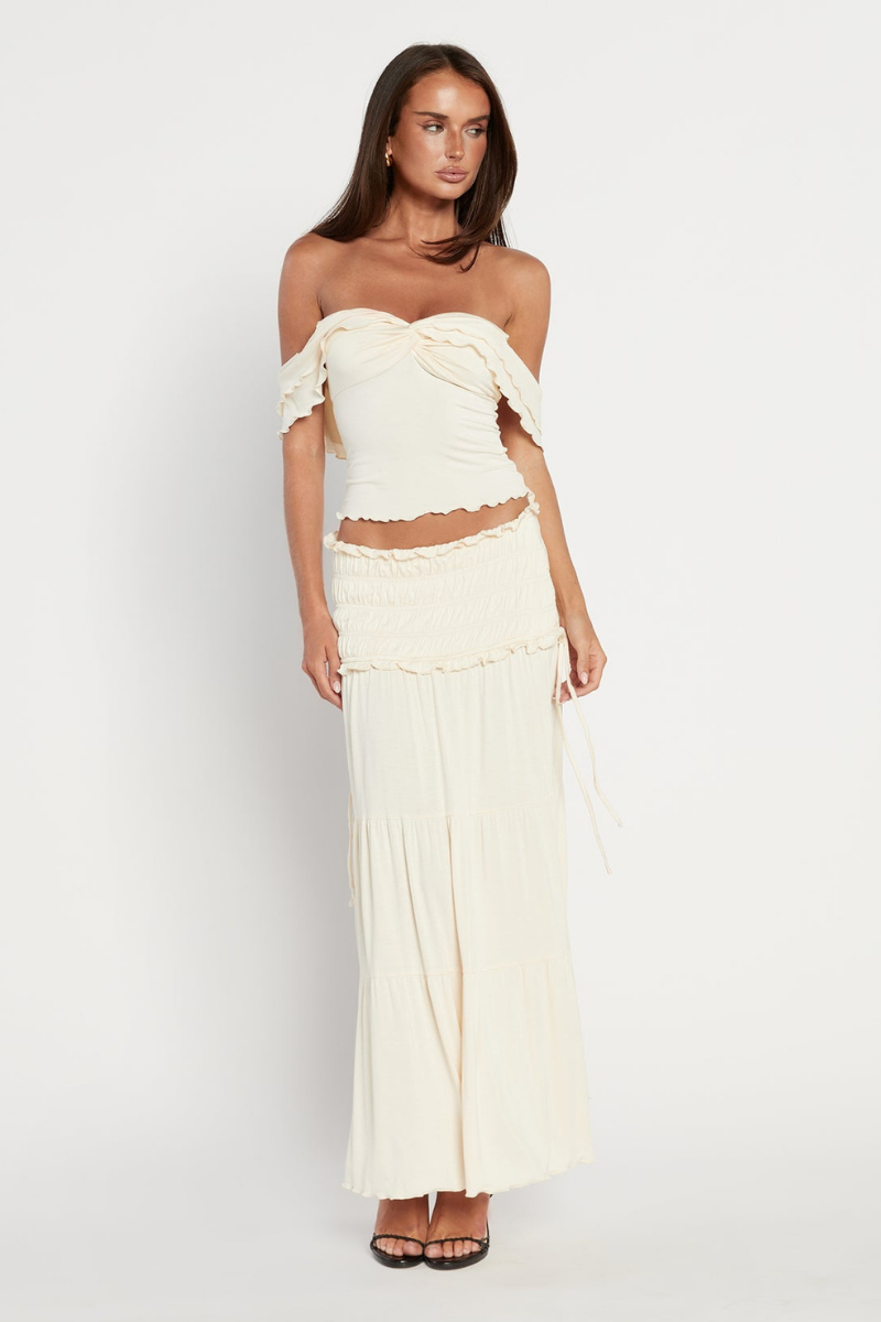 Apricot - A woman wearing a cream-coloured two-piece set featuring an off-the-shoulder top with a twisted bust and a smocked, tiered maxi skirt with ruffle trims.