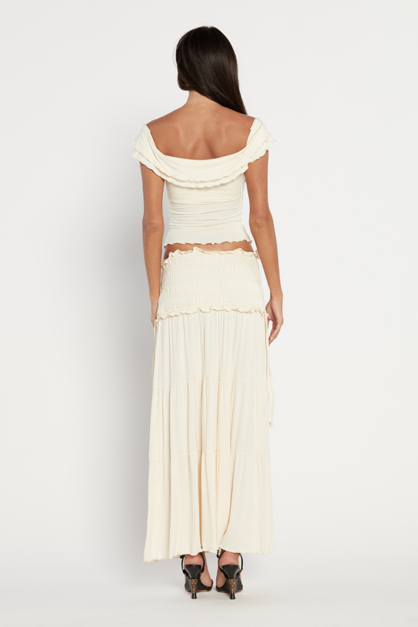 Apricot - A woman wearing a cream-coloured two-piece set featuring an off-the-shoulder top with a twisted bust and a smocked, tiered maxi skirt with ruffle trims.