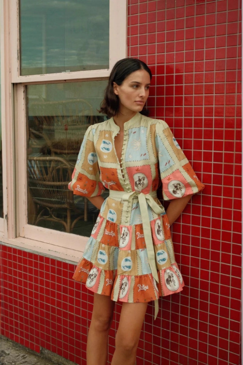 A woman wearing a short, colourful patchwork-print mini dress featuring a band collar, button-front bodice, puff sleeves, a tie waist, and a tiered skirt.