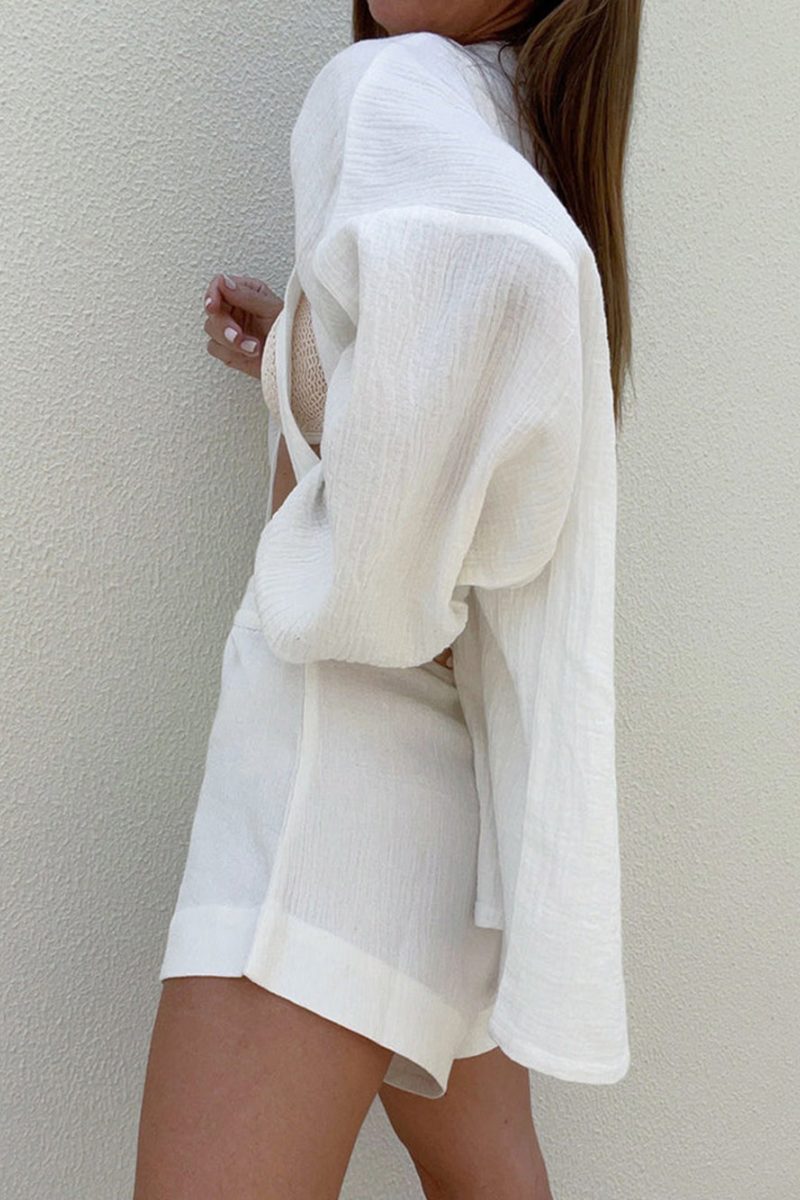 White - Woman wearing a white, crinkled-texture wrap playsuit with voluminous sleeves and a tie waist, holding a brown clutch bag.