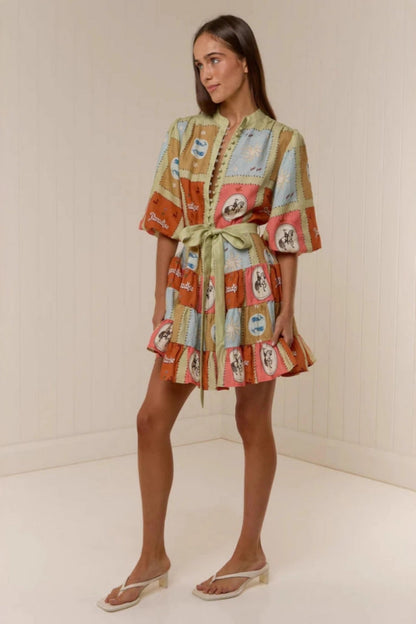 A woman wearing a short, colourful patchwork-print mini dress featuring a band collar, button-front bodice, puff sleeves, a tie waist, and a tiered skirt.