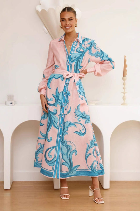 Green - Full-length view of a woman wearing a pink maxi shirt dress featuring a bold blue baroque print, long blouson sleeves, a collared neck, and a matching tie-belt at the waist.