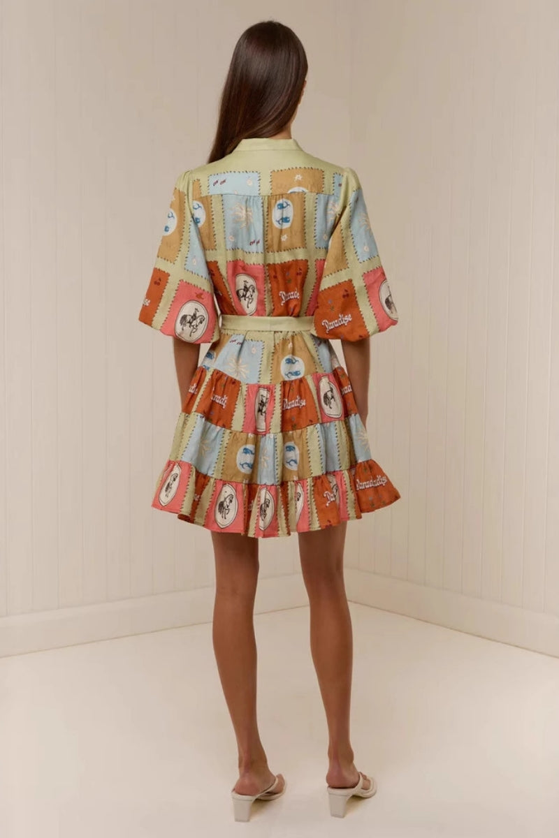 A woman wearing a short, colourful patchwork-print mini dress featuring a band collar, button-front bodice, puff sleeves, a tie waist, and a tiered skirt.