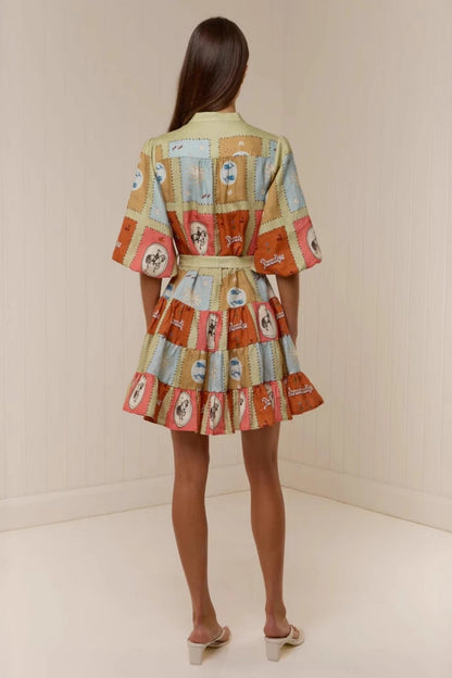 A woman wearing a short, colourful patchwork-print mini dress featuring a band collar, button-front bodice, puff sleeves, a tie waist, and a tiered skirt.