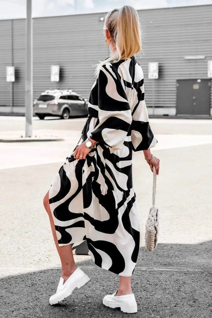 Black - A woman wearing a long, black and white abstract print shirt dress with a tie waist, blouson sleeves, and a high side slit, paired with white chunky loafers.
