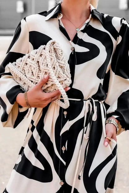 Black - A woman wearing a long, black and white abstract print shirt dress with a tie waist, blouson sleeves, and a high side slit, paired with white chunky loafers.