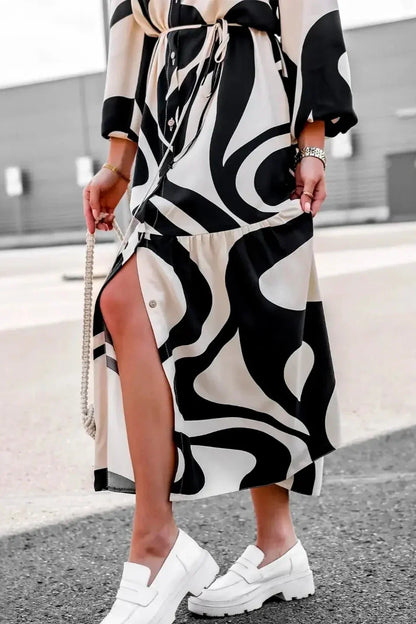 Black - A woman wearing a long, black and white abstract print shirt dress with a tie waist, blouson sleeves, and a high side slit, paired with white chunky loafers.