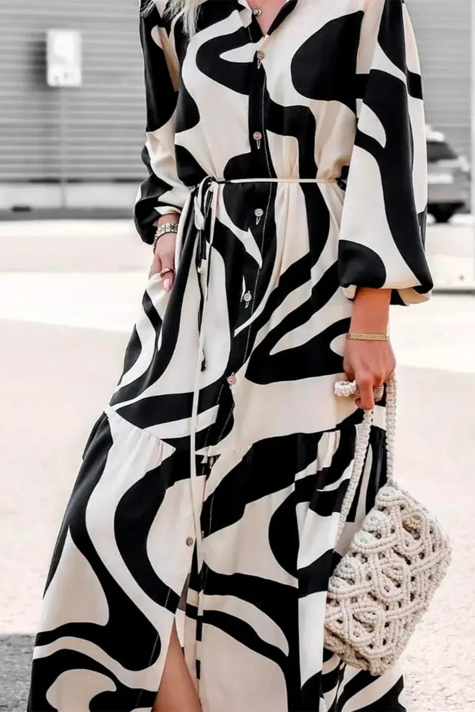 Black - A woman wearing a long, black and white abstract print shirt dress with a tie waist, blouson sleeves, and a high side slit, paired with white chunky loafers.