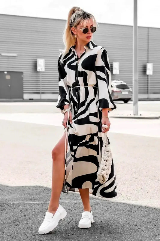 Black - A woman wearing a long, black and white abstract print shirt dress with a tie waist, blouson sleeves, and a high side slit, paired with white chunky loafers.