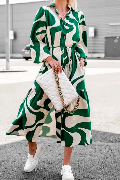 Green - A woman wearing a long, black and white abstract print shirt dress with a tie waist, blouson sleeves, and a high side slit, paired with white chunky loafers.