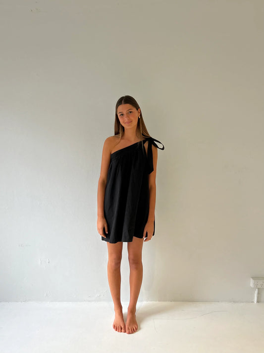 A woman wearing a black, one-shoulder mini dress with a prominent tie detail on the strap and a loose, voluminous trapeze silhouette, standing against a plain white wall.