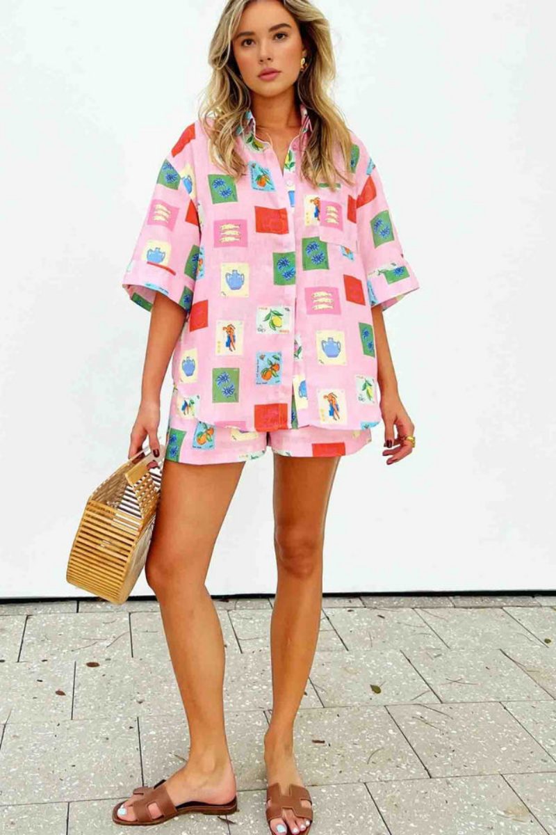 Pink - Women's pink short-sleeve belted playsuit featuring a colourful Mediterranean novelty print with oranges and vases, styled with sunglasses and a bamboo bag.