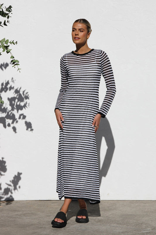 Woman wearing a long-sleeve, black and white striped crochet maxi dress with a crew neckline, standing outdoors against a white wall.