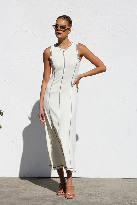 A woman stands against a white wall wearing a sleeveless, form-fitting, off-white midi dress. The dress has a high neckline and is detailed with dark vertical contrast stitching along the seams and armholes.