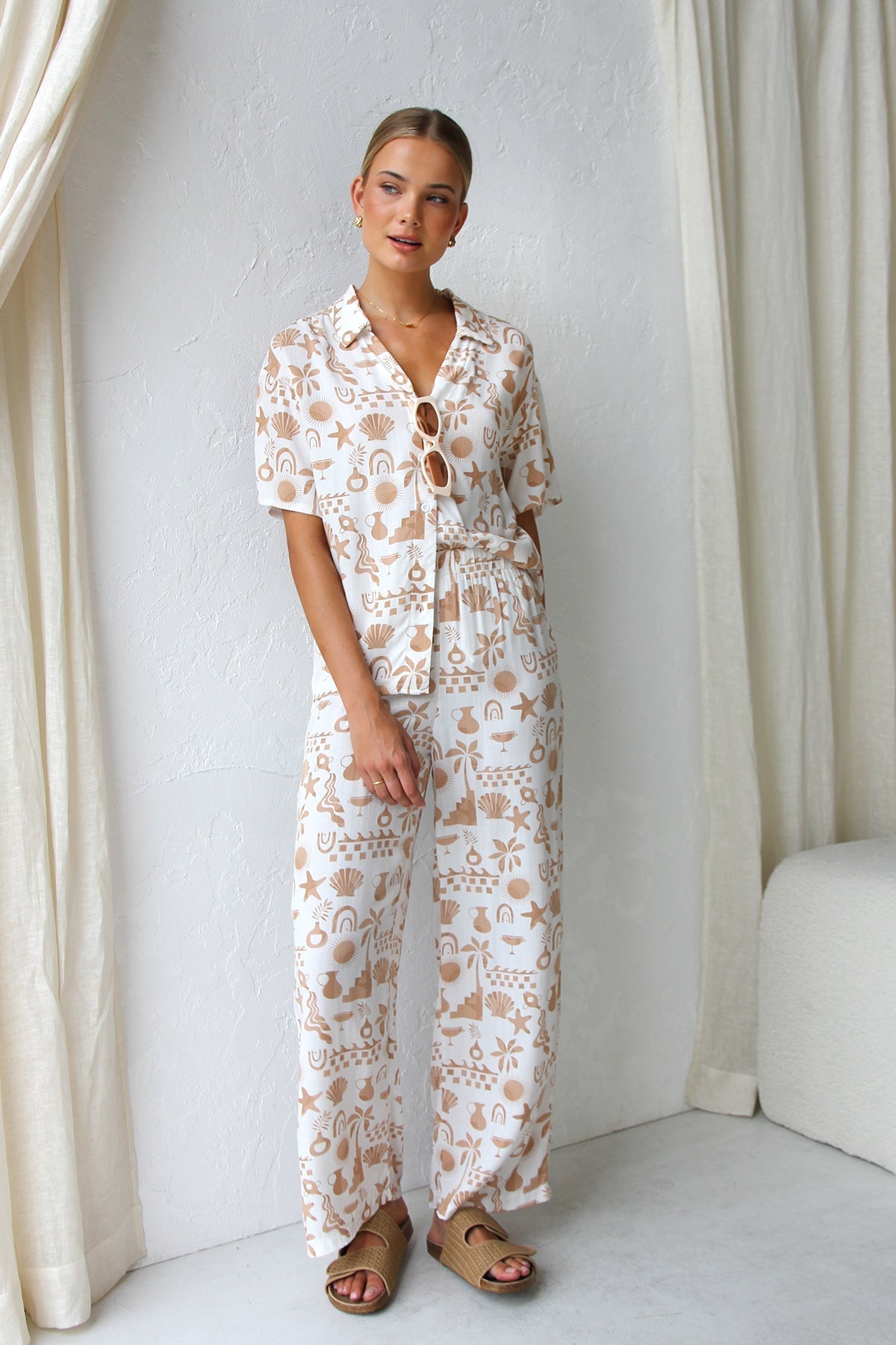 A woman modelling a relaxed-fit, wide-leg trouser and matching short-sleeve shirt co-ord set, featuring a beige coastal vacation print on a white background.