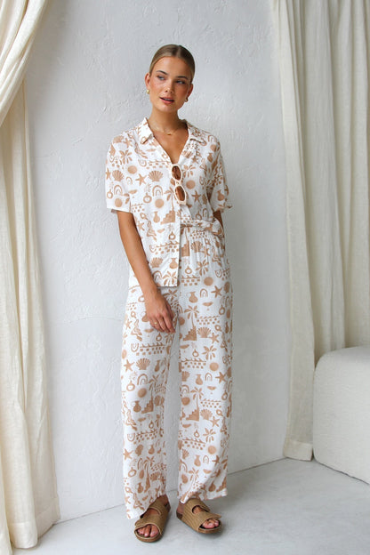 A woman modelling a relaxed-fit, wide-leg trouser and matching short-sleeve shirt co-ord set, featuring a beige coastal vacation print on a white background.