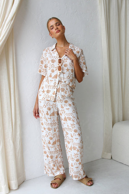 Multicolor - A woman modelling a relaxed-fit, wide-leg trouser and matching short-sleeve shirt co-ord set, featuring a beige coastal vacation print on a white background.