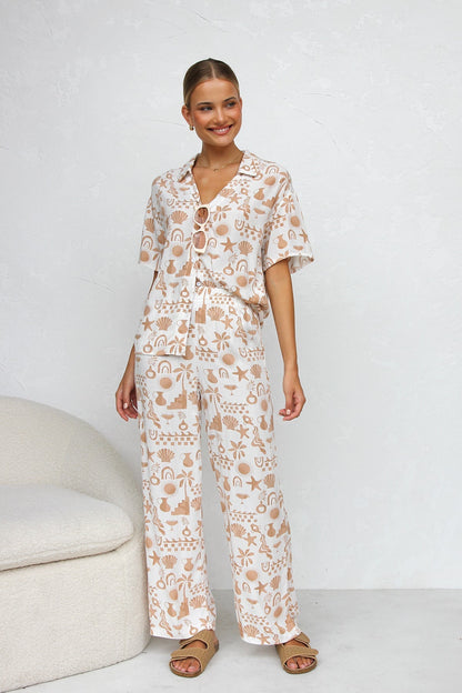 A woman modelling a relaxed-fit, wide-leg trouser and matching short-sleeve shirt co-ord set, featuring a beige coastal vacation print on a white background.