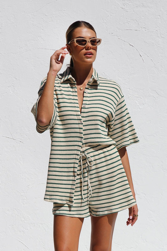 A woman wearing a matching cream and green horizontally striped two-piece set, featuring a short-sleeved button-down collared shirt and drawstring shorts, standing against a white textured wall.