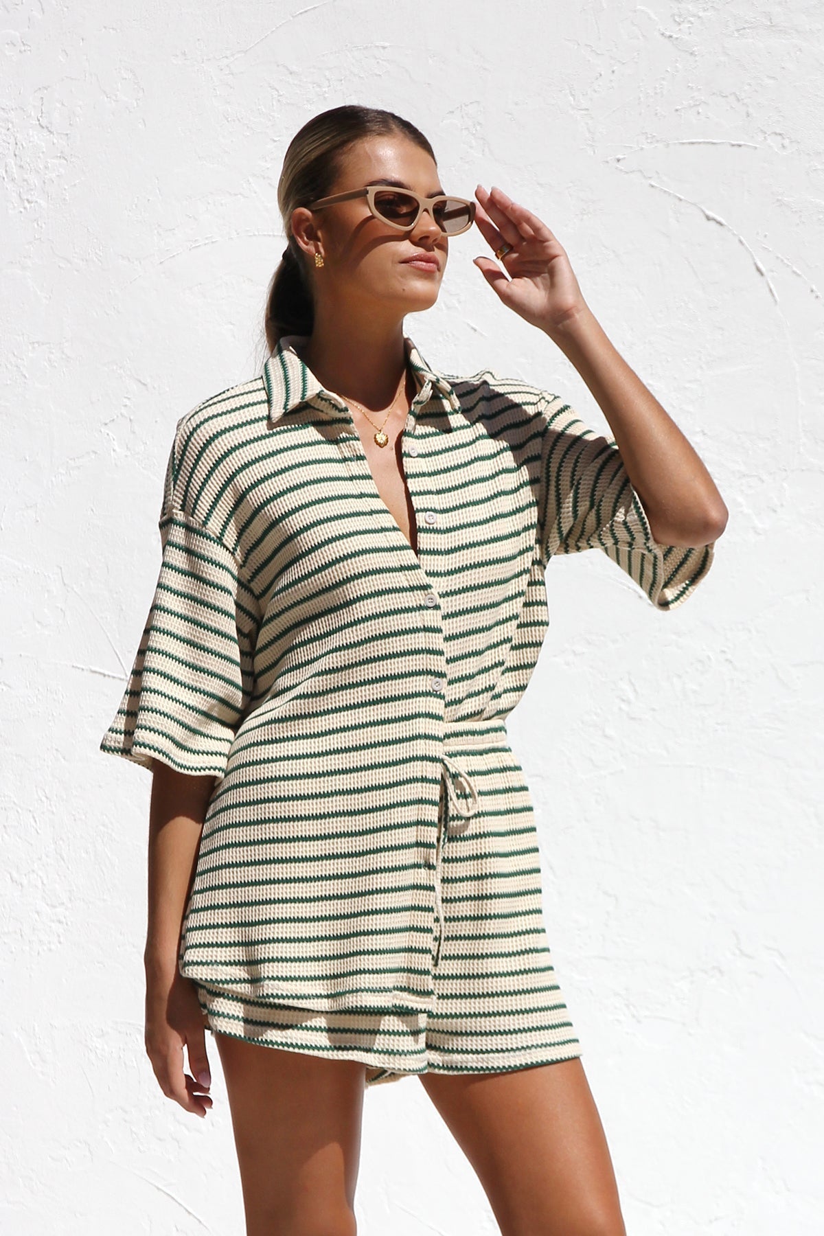 A woman wearing a matching cream and green horizontally striped two-piece set, featuring a short-sleeved button-down collared shirt and drawstring shorts, standing against a white textured wall.