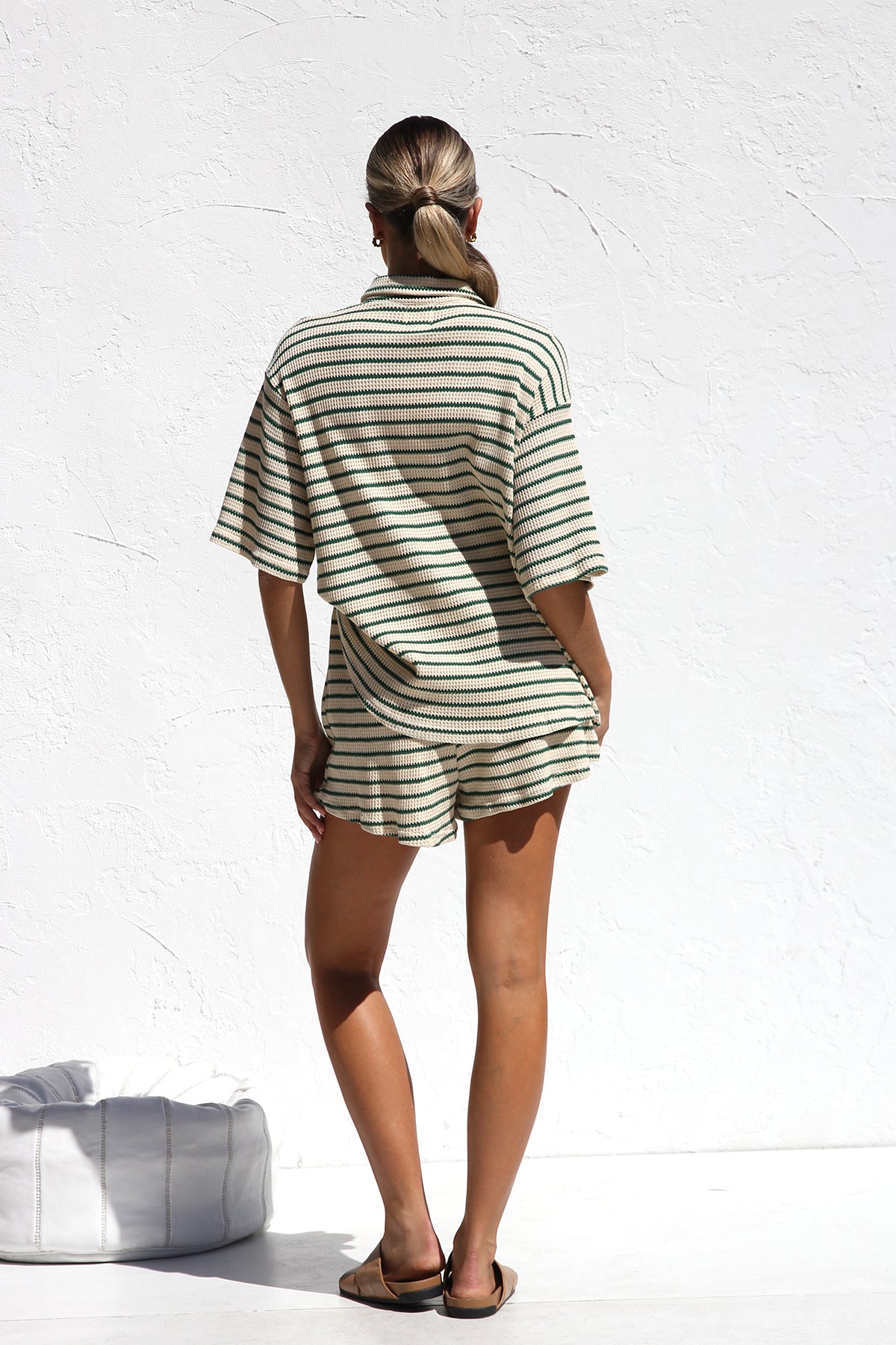A woman wearing a matching cream and green horizontally striped two-piece set, featuring a short-sleeved button-down collared shirt and drawstring shorts, standing against a white textured wall.
