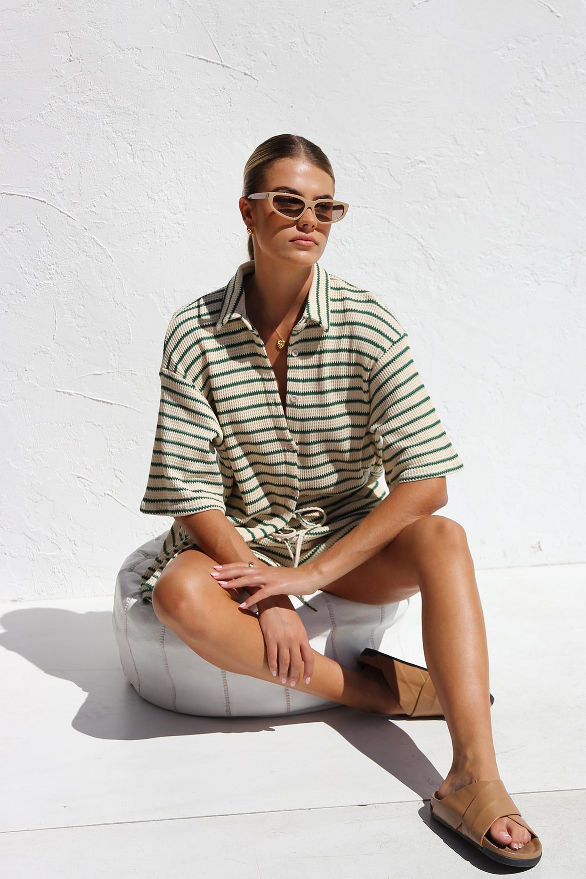 A woman wearing a matching cream and green horizontally striped two-piece set, featuring a short-sleeved button-down collared shirt and drawstring shorts, standing against a white textured wall.