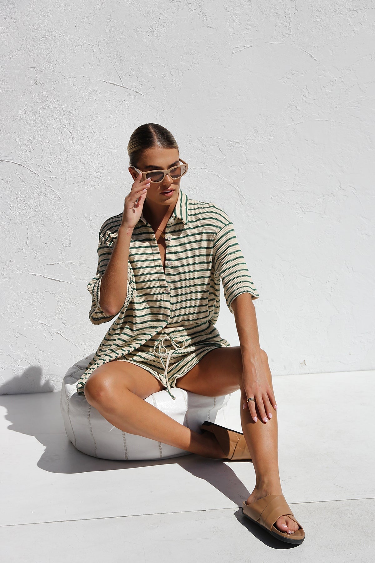 A woman wearing a matching cream and green horizontally striped two-piece set, featuring a short-sleeved button-down collared shirt and drawstring shorts, standing against a white textured wall.