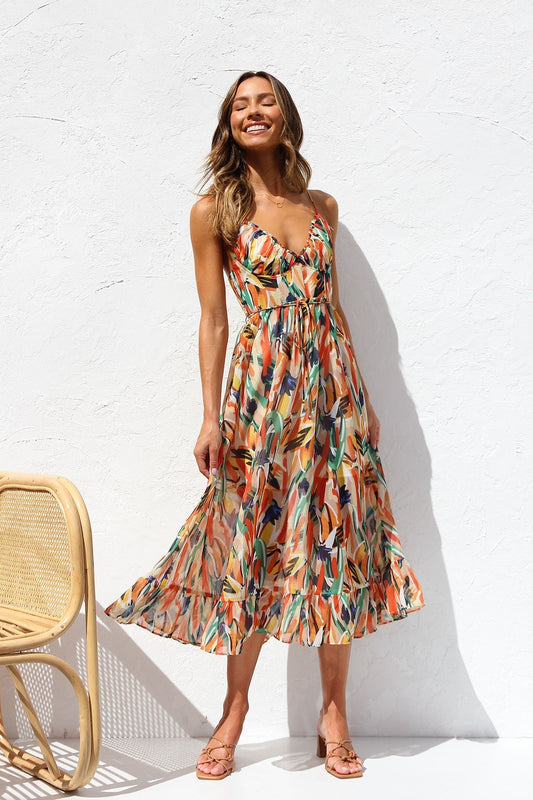 A woman smiling outdoors wearing a vibrant, abstract brushstroke print midi dress featuring a V-neckline, thin straps, a tie waist, and a ruffled hem.