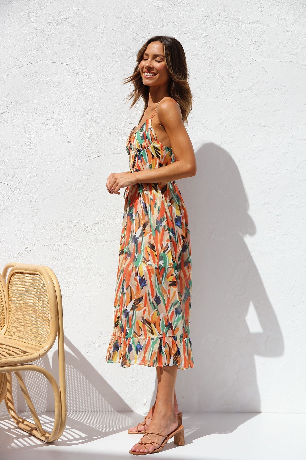 Multicolor - A woman smiling outdoors wearing a vibrant, abstract brushstroke print midi dress featuring a V-neckline, thin straps, a tie waist, and a ruffled hem.