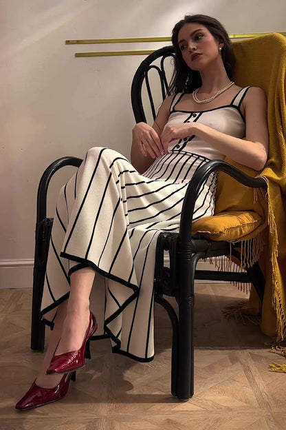 White - Woman wearing an elegant off-white midi knit dress with black contrast binding on the square neckline and straps, a buttoned bodice, and a flared A-line skirt featuring black vertical stripes.