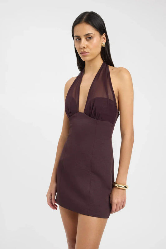 A woman modelling a dark plum, form-fitting mini dress featuring a structured corset-style bodice, a plunging sheer halter neckline, and tailored vertical seams.