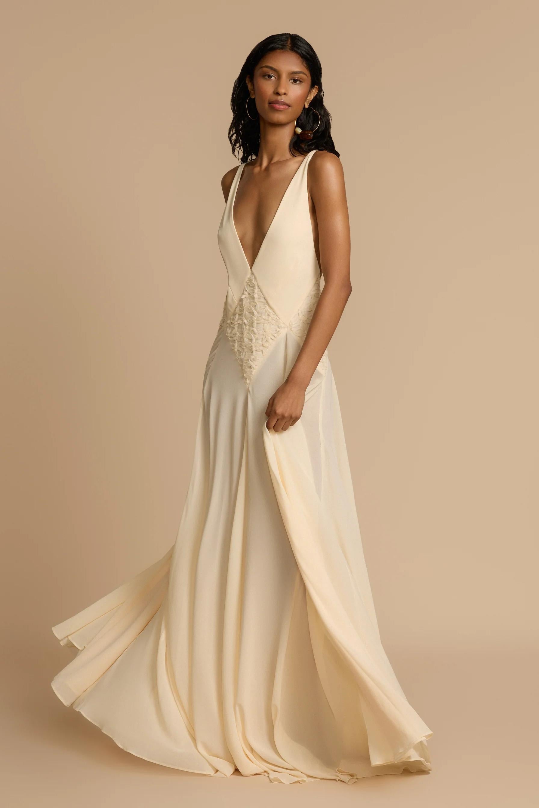 Ivory - A woman wearing an elegant, flowing off-white maxi dress featuring a deep plunging V-neckline and a textured appliqué waist panel, standing against a neutral background.