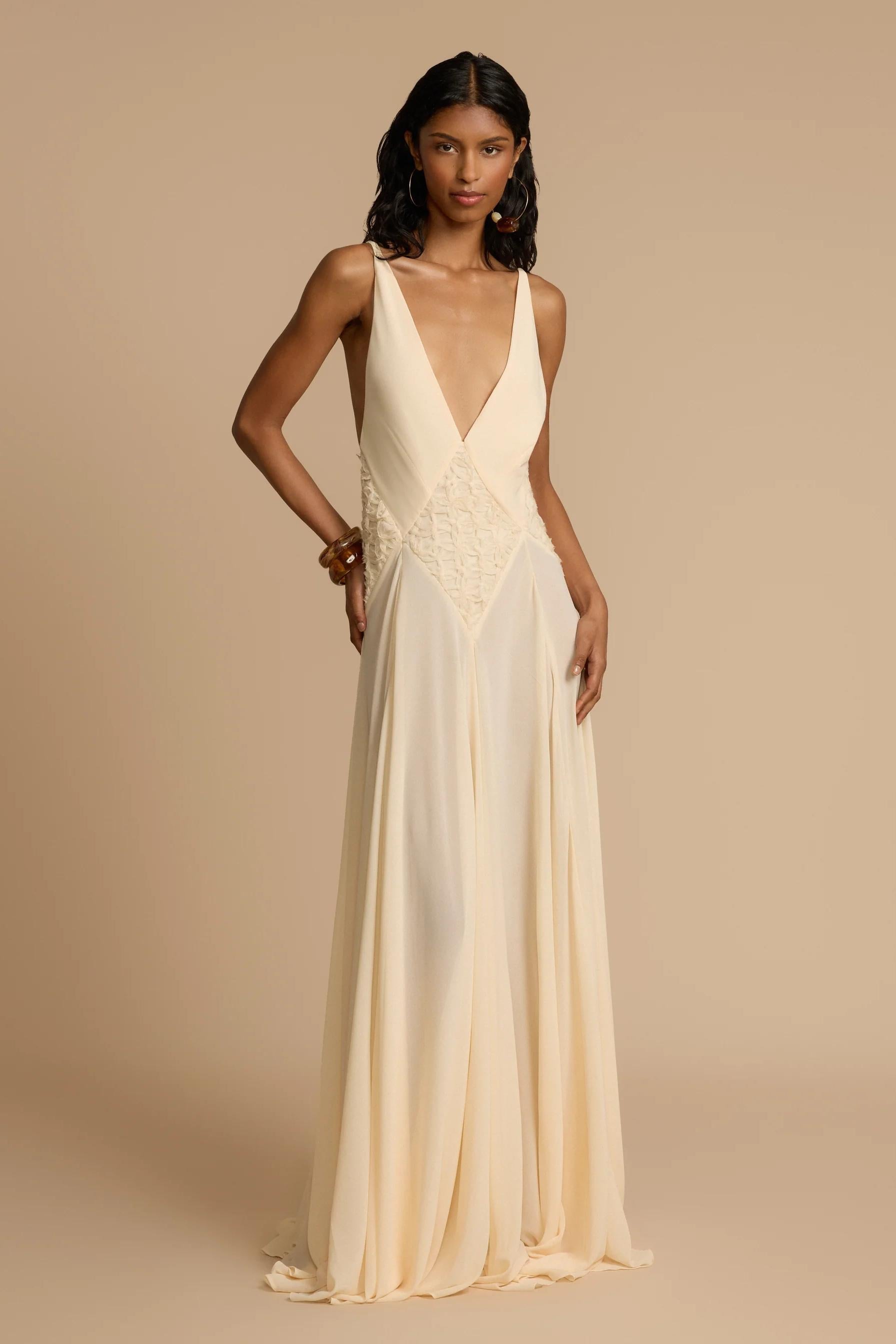Ivory - A woman wearing an elegant, flowing off-white maxi dress featuring a deep plunging V-neckline and a textured appliqué waist panel, standing against a neutral background.