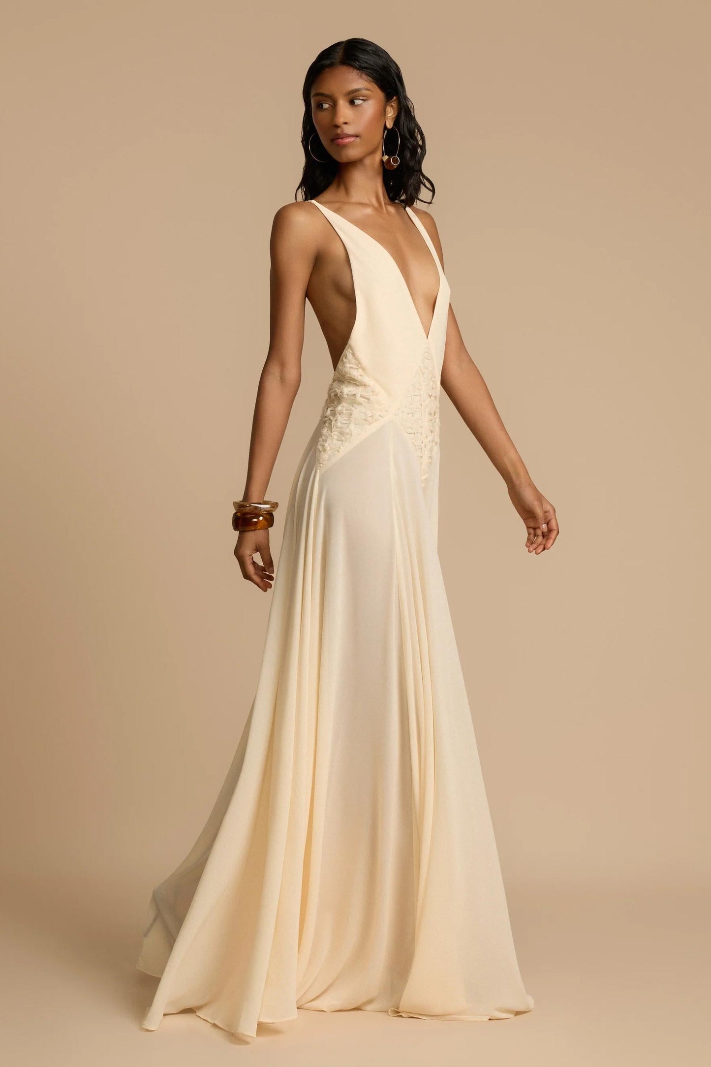 Ivory - A woman wearing an elegant, flowing off-white maxi dress featuring a deep plunging V-neckline and a textured appliqué waist panel, standing against a neutral background.
