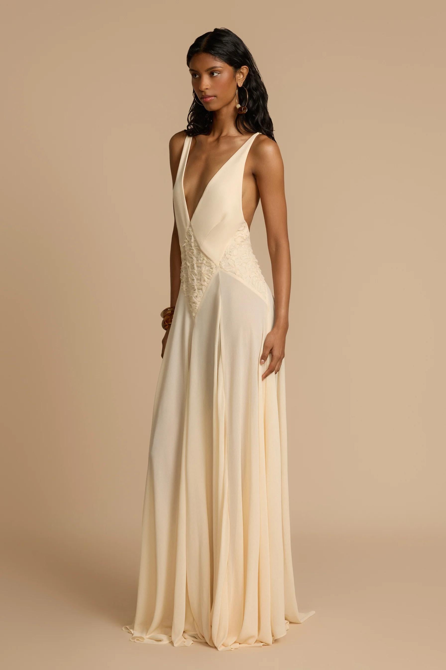 Ivory - A woman wearing an elegant, flowing off-white maxi dress featuring a deep plunging V-neckline and a textured appliqué waist panel, standing against a neutral background.