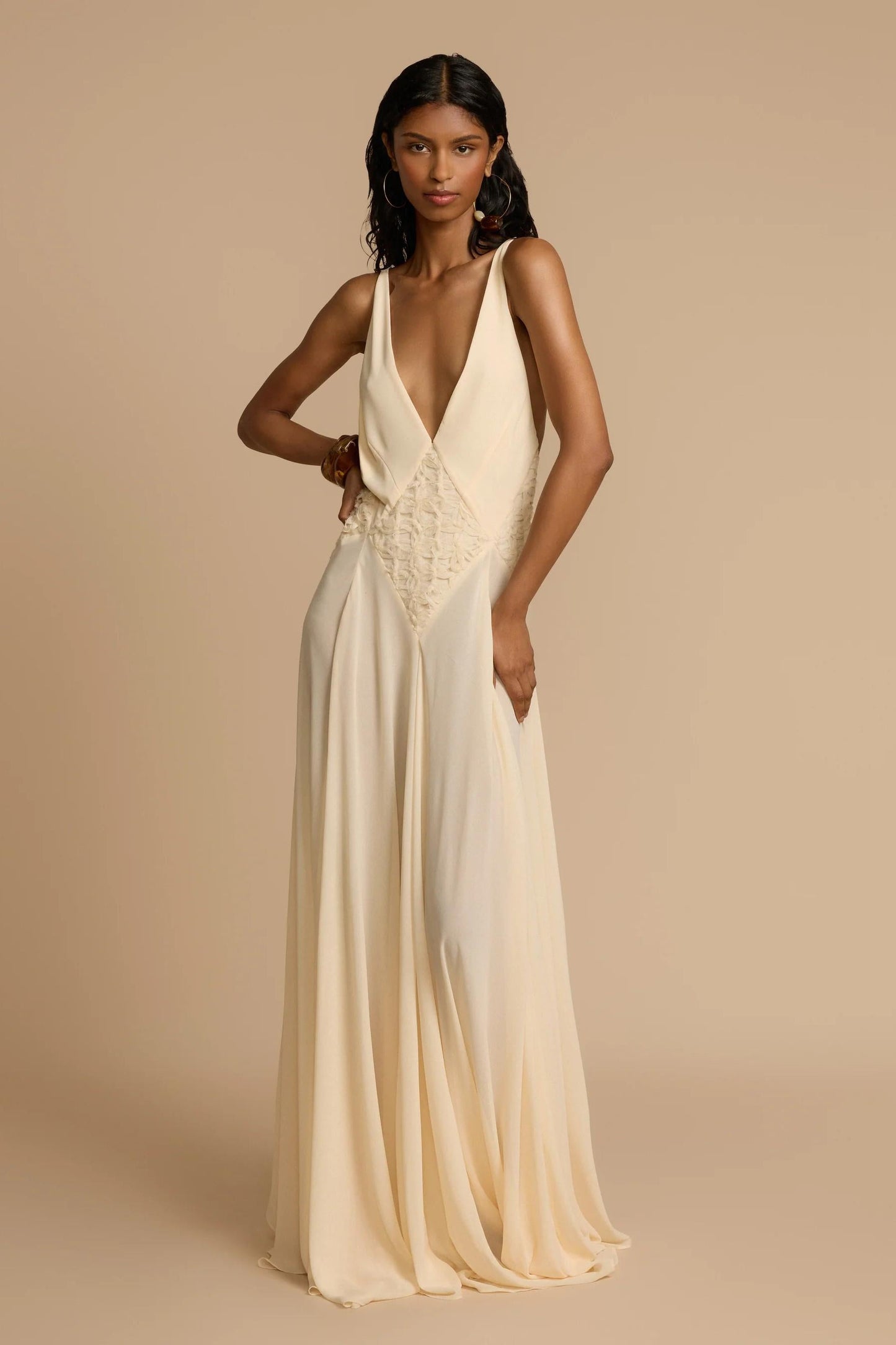 Ivory - A woman wearing an elegant, flowing off-white maxi dress featuring a deep plunging V-neckline and a textured appliqué waist panel, standing against a neutral background.