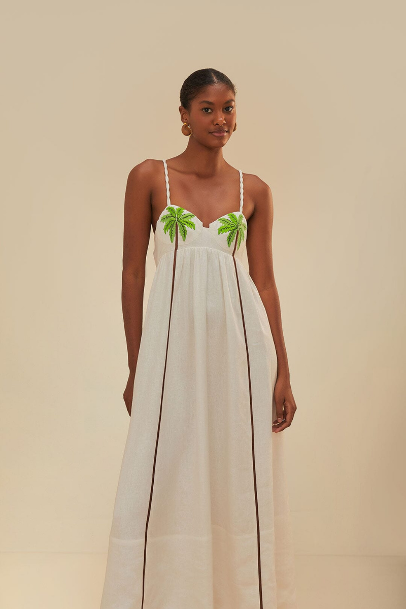 White - A woman wearing a flowing white maxi dress with braided spaghetti straps, a sweetheart neckline featuring embroidered green palm trees on the cups, and long brown vertical stripes down the skirt.