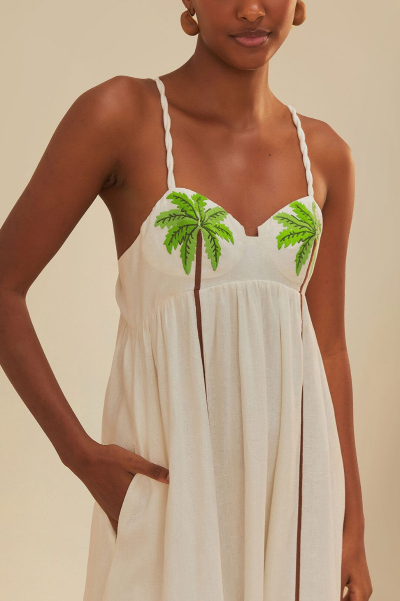 White - A woman wearing a flowing white maxi dress with braided spaghetti straps, a sweetheart neckline featuring embroidered green palm trees on the cups, and long brown vertical stripes down the skirt.