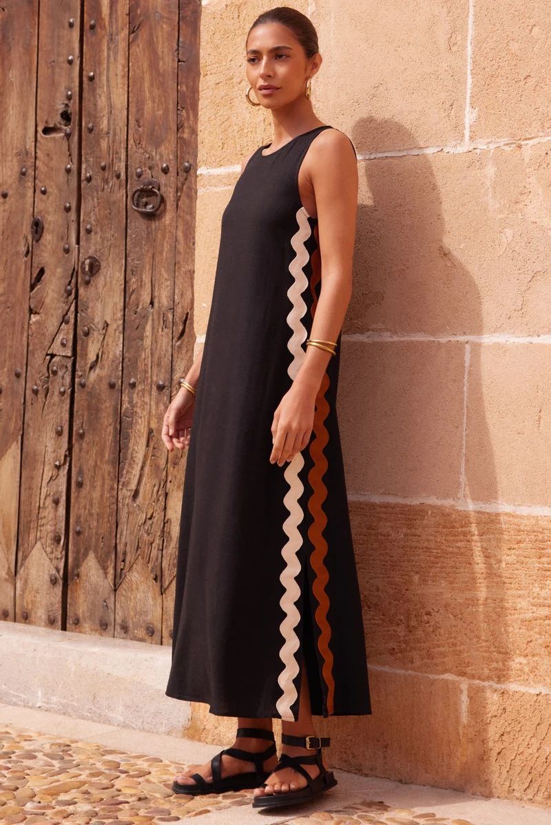 Black - Full-length view of a woman in a wide-brimmed hat and a sleeveless, rust-orange midi shift dress featuring a white wavy trim down the side. She stands barefoot against a peach wall with blue shutters.