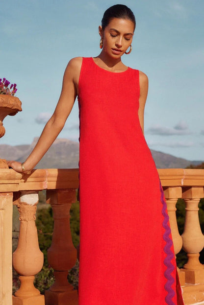 Brown - Full-length view of a woman in a wide-brimmed hat and a sleeveless, rust-orange midi shift dress featuring a white wavy trim down the side. She stands barefoot against a peach wall with blue shutters.