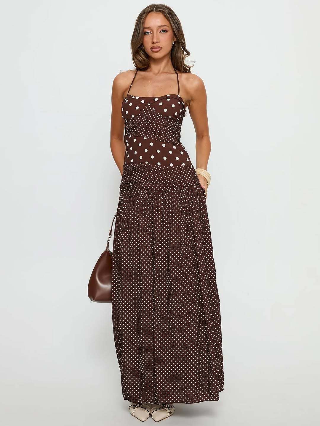 Cocoa - A woman wearing a brown maxi dress featuring a mixed white polka dot print, a halter neckline, a fitted panelled bodice, and a flowing gathered skirt.