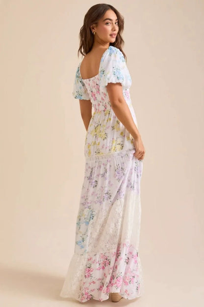 Colorful - A woman wearing a white, tiered maxi dress with a smocked bodice, short puff sleeves, and a pastel patchwork floral print with lace inserts.